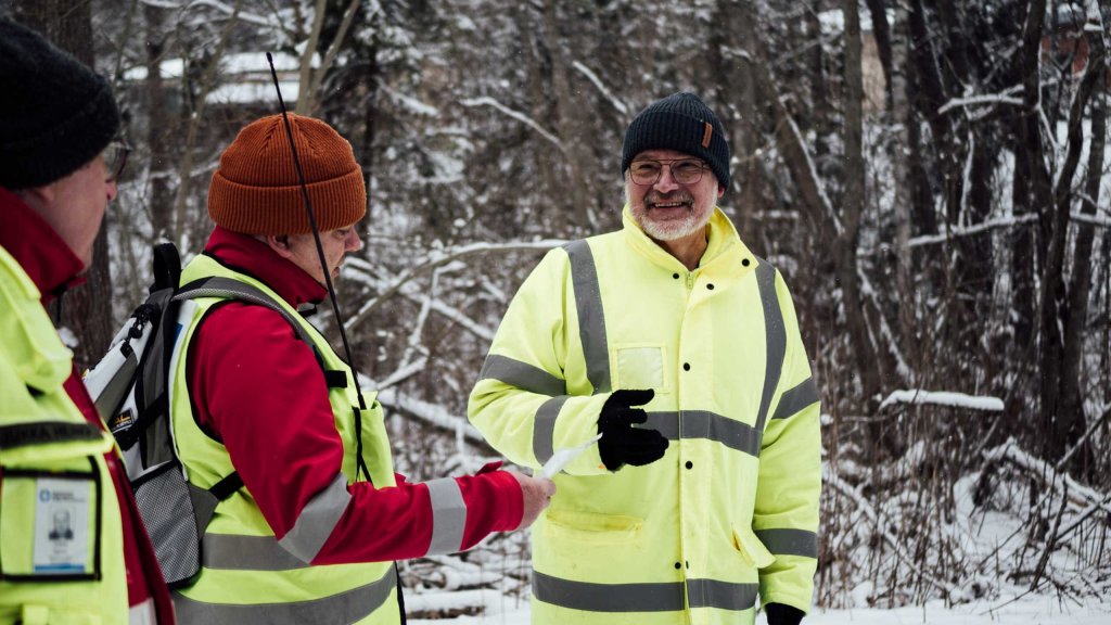 Kaksi vapaaehtoista kuuntelevat, kun yksi juttelee heille hymyillen. Taustalla talvinen maisema.